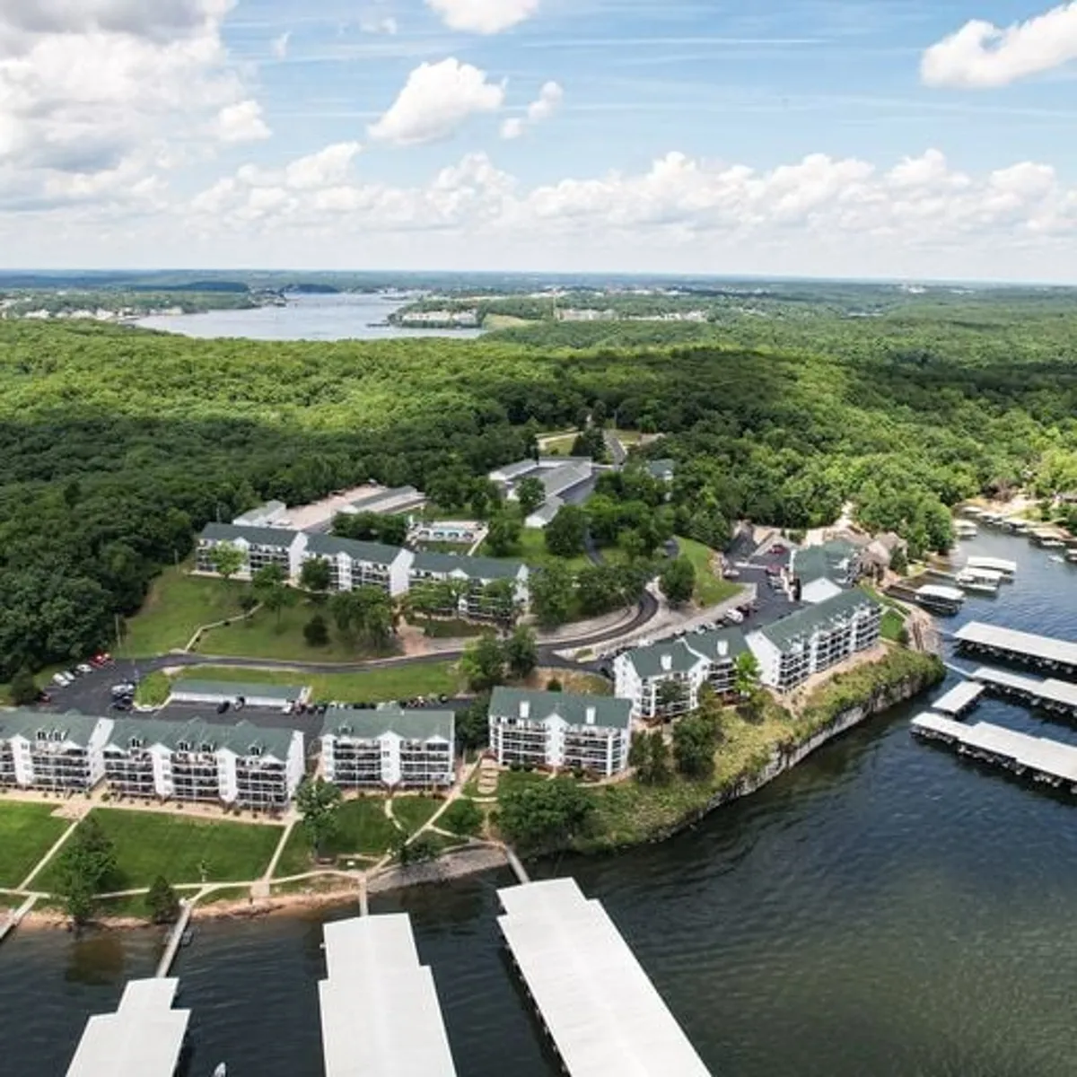 A tranquil lakeside setting showing the expansive lake, marina with boat slips, surrounding forests, residential buildings, and walking pathways. Activities like boating, fishing, hiking, and picnicking are visible or implied by the landscape and amenities. The scene is bathed in the warm light of sunset, enhancing the peaceful mood.