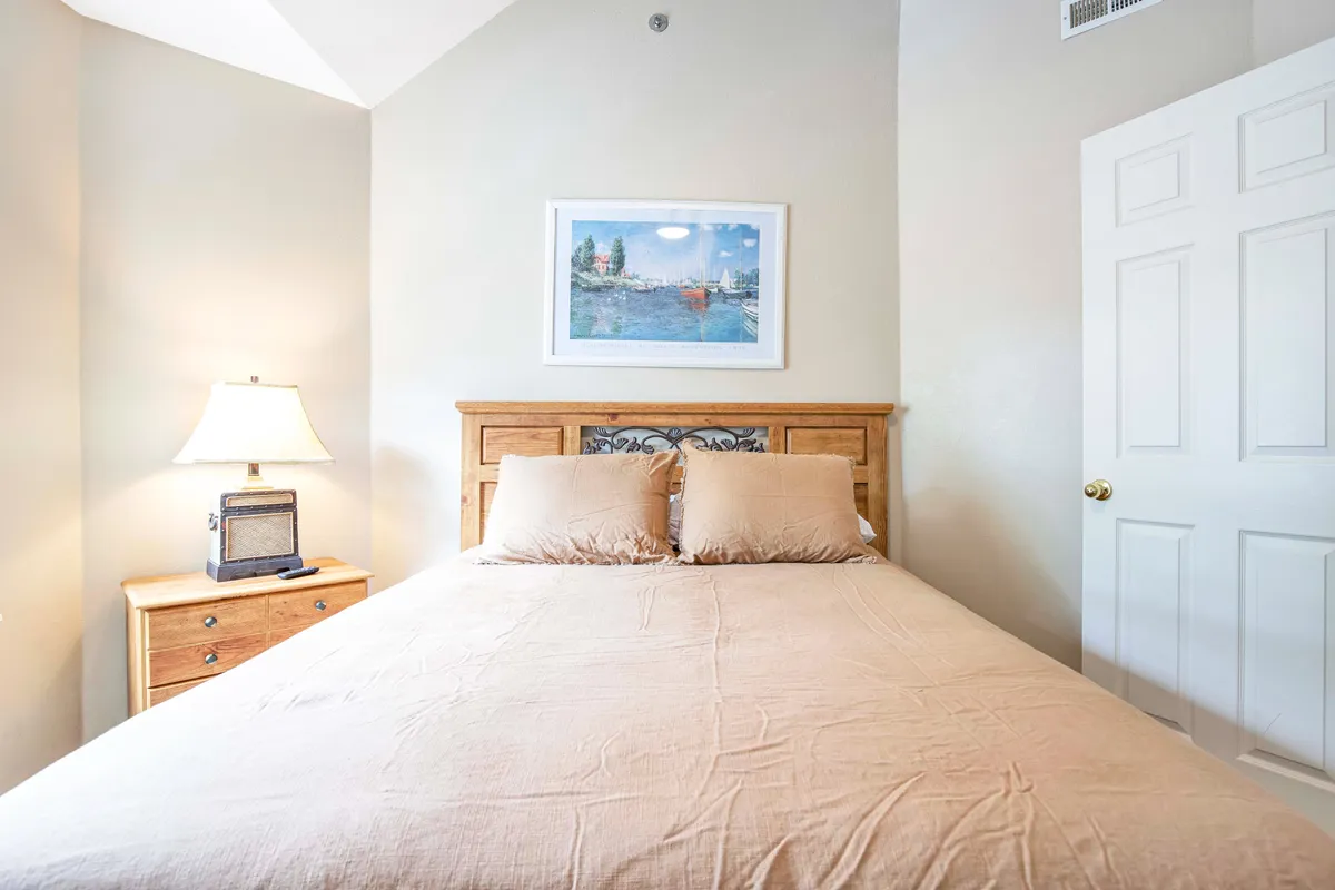 A tranquil bedroom featuring a bed with neutral bedding, soft natural light streaming through a nearby window, a piece of calming artwork above the bed, and a table lamp on the nightstand. The mood is peaceful and inviting, with an uncluttered design emphasizing comfort and relaxation.