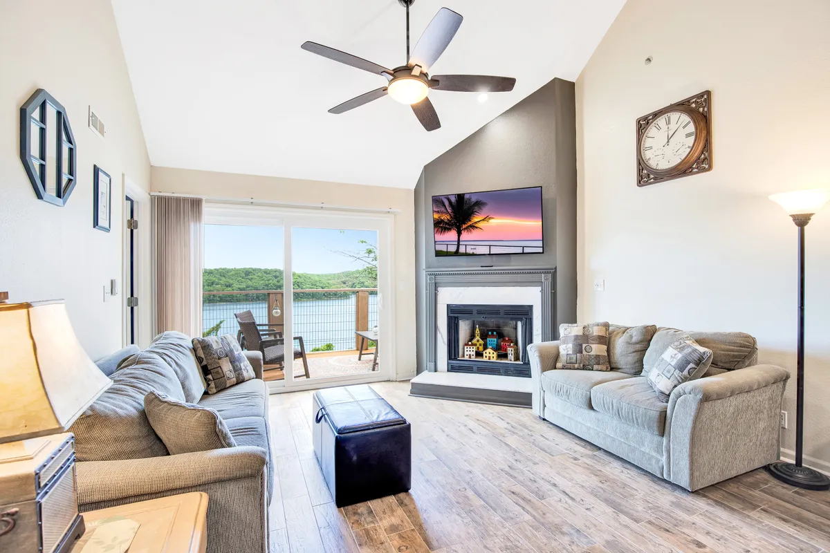 A spacious living area featuring large windows that offer sweeping views of the lake, a cozy fireplace, a TV displaying a sunset, and a comfortable seating arrangement. The room is filled with natural light, inviting both relaxation and social gatherings, while the water view establishes a direct connection to the surrounding landscape.