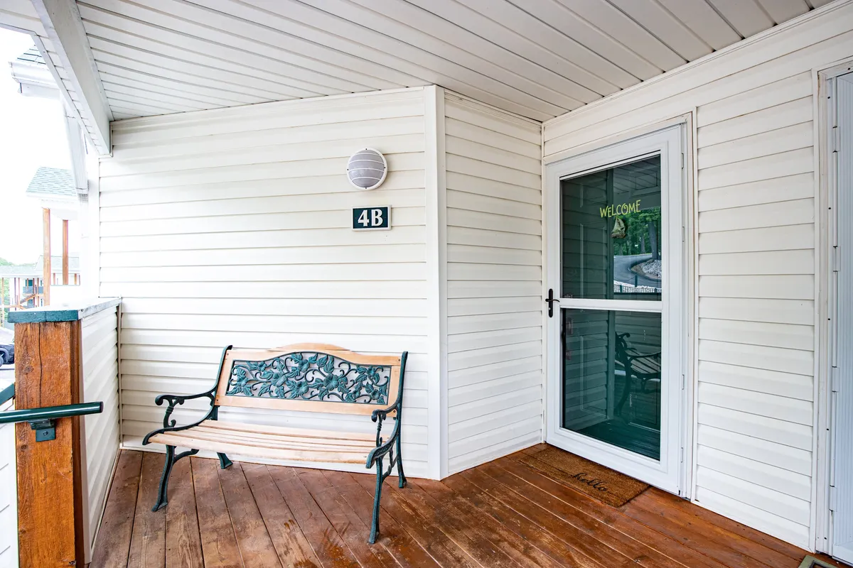 The condo entrance with a decorative bench, welcoming sign, and front door, framed by simple landscaping. The scene conveys a warm first impression and a comforting sense of arrival home, suggesting both hospitality and privacy.