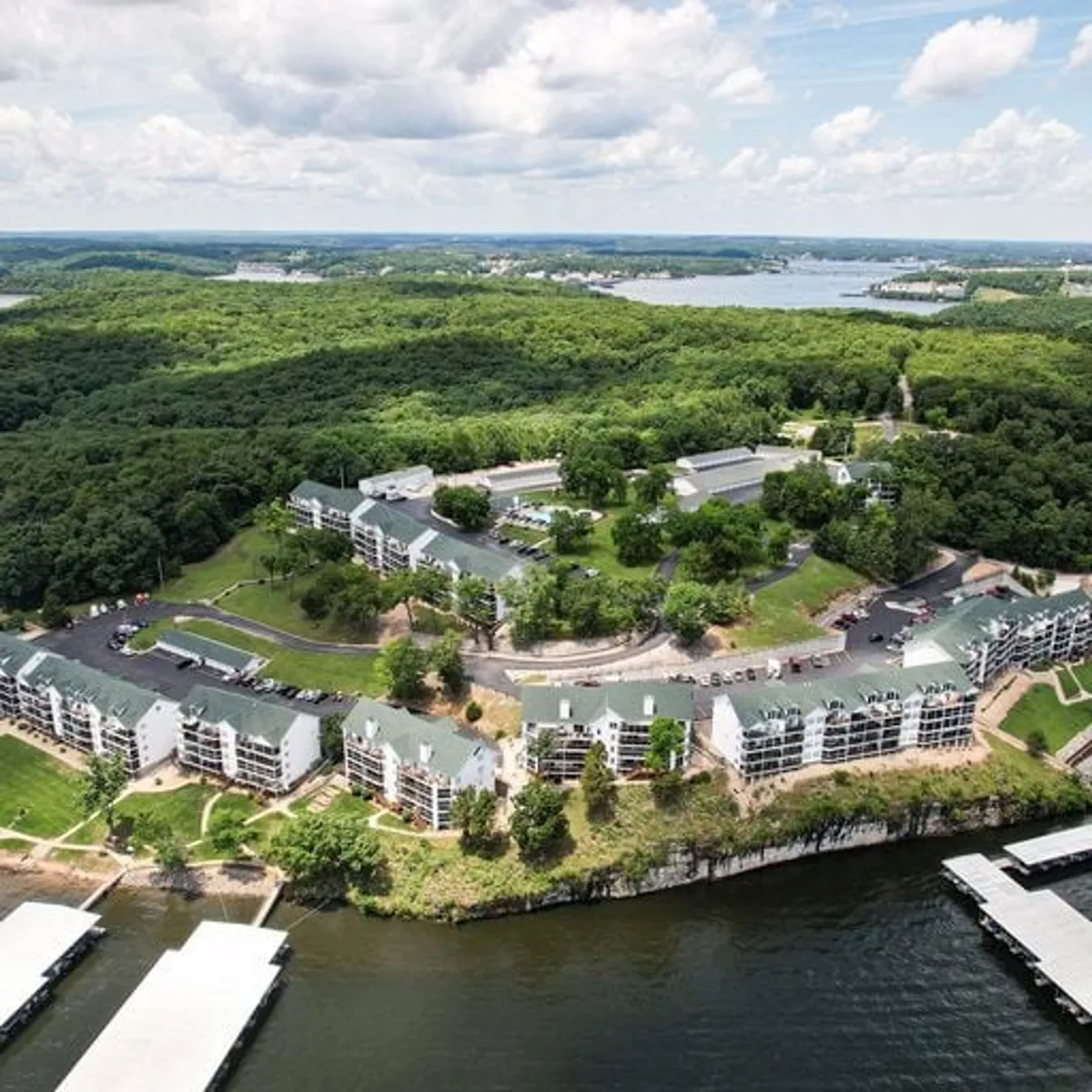 A waterfront view with a dock extending into the lake, surrounded by lush green landscape and buildings with green roofs. The scene invites boating, fishing, picnicking, and other recreational activities, with the calm water reflecting the sky and shoreline.