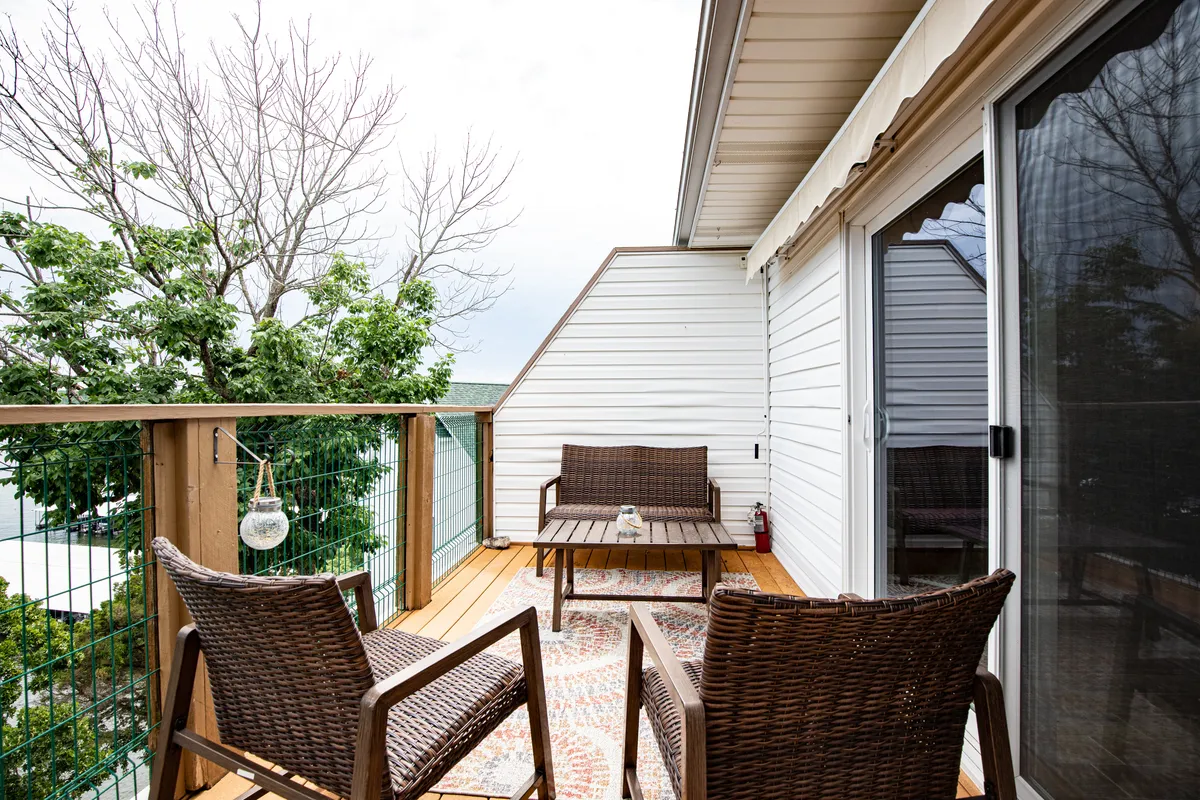 A serene waterfront scene featuring wicker chairs and a wooden table set on an outdoor patio, surrounded by lush green foliage and offering a peaceful view of the water. The inviting setup suggests a perfect spot for relaxing evenings, casual gatherings, or enjoying a drink while watching the sunset.