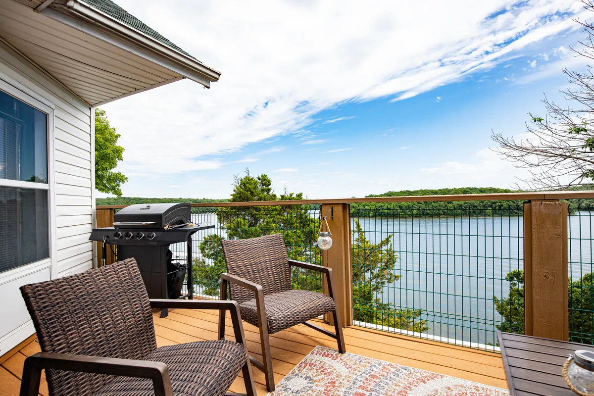 This image showcases a top-floor deck complete with outdoor seating and a grill, overlooking the tranquil lake. The view extends across the sparkling water, framed by lush green trees, and invites you to enjoy both sunrise and sunset in a serene and private alfresco setting.