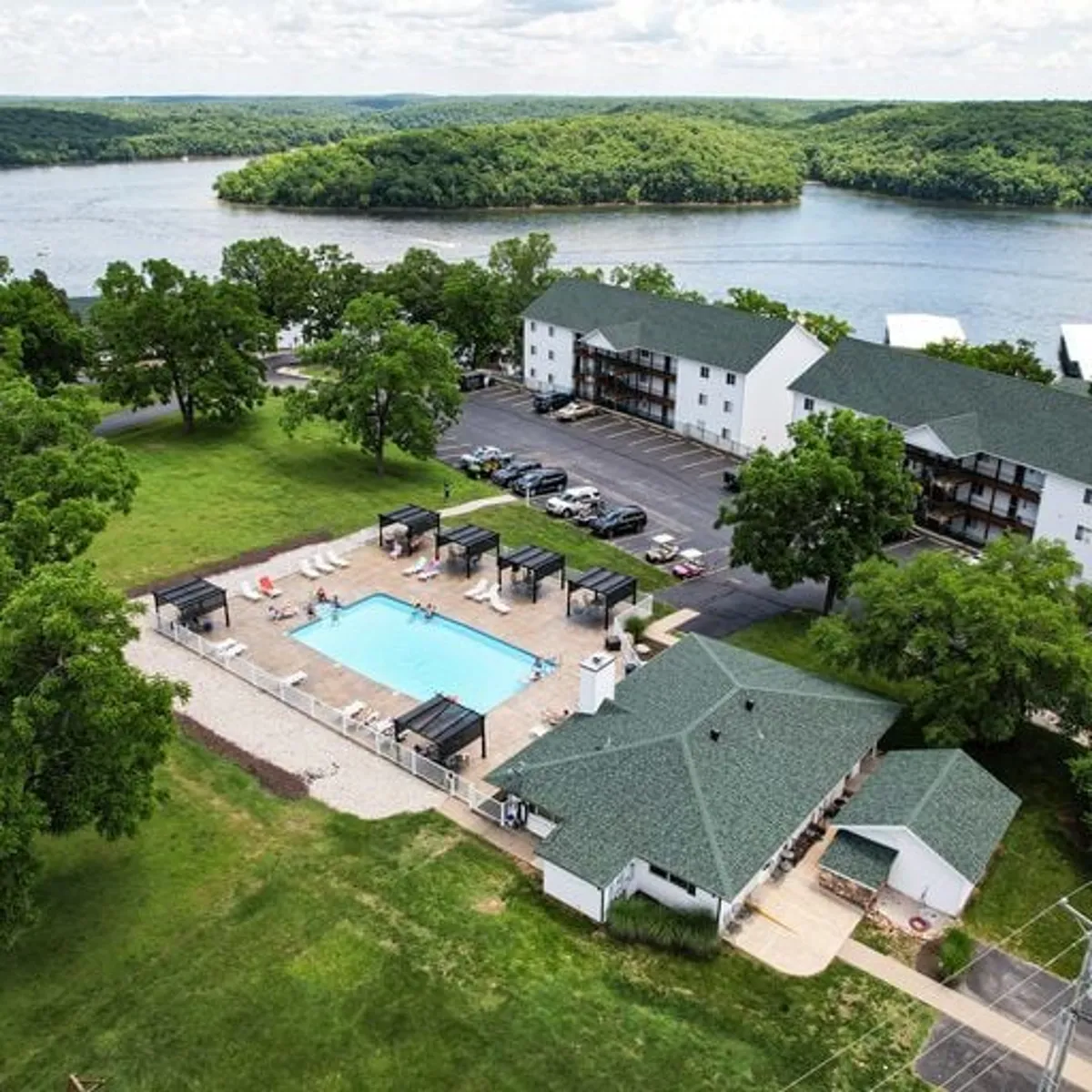 The photo features a sparkling swimming pool surrounded by lounge chairs, with a beautifully landscaped green area and a picturesque waterfront in the background. The setting is serene yet lively, ideal for both adults relaxing and children playing nearby.