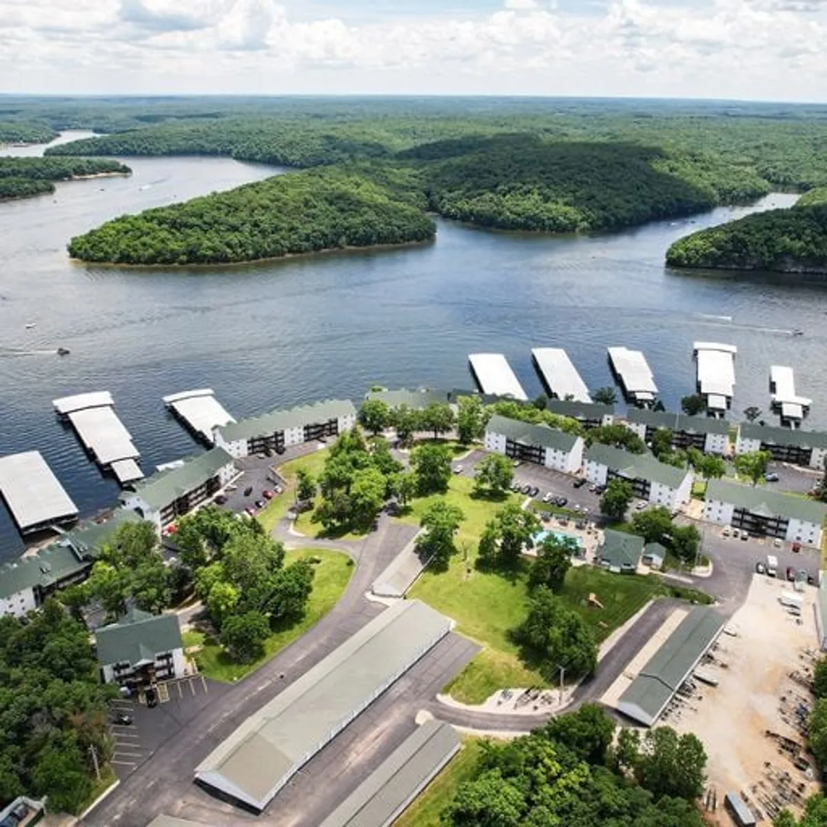 A vibrant view of the marina, boat docks, swimming pool area, surrounding green spaces, and the shimmering water of the lake bordered by wooded hills. The overall atmosphere is one of recreation and relaxation, with the sunset casting a warm glow over the landscape.