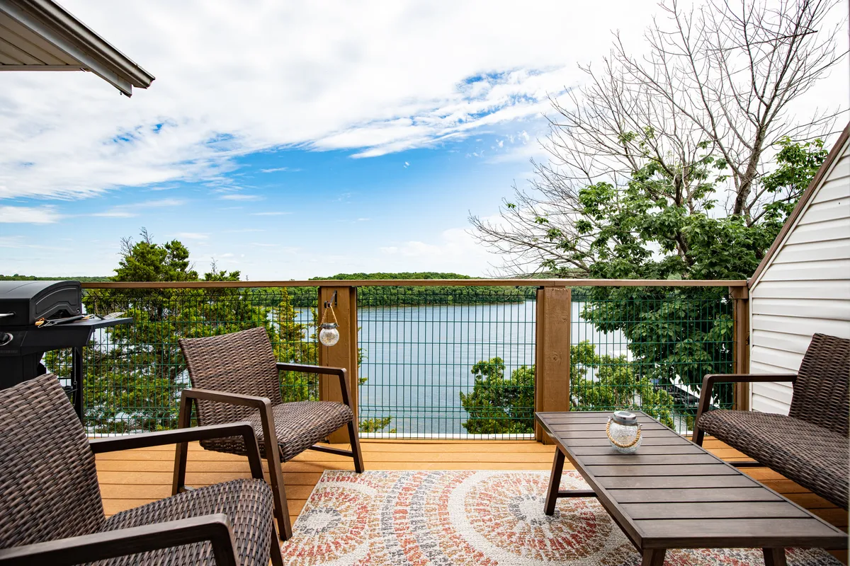 The image showcases a serene lake view from the condo's balcony, with comfortable outdoor seating arranged on a decorative rug. A grill is nearby, inviting outdoor dining and barbecues, while the atmosphere suggests relaxation, socializing, and enjoying nature—especially during sunset.
