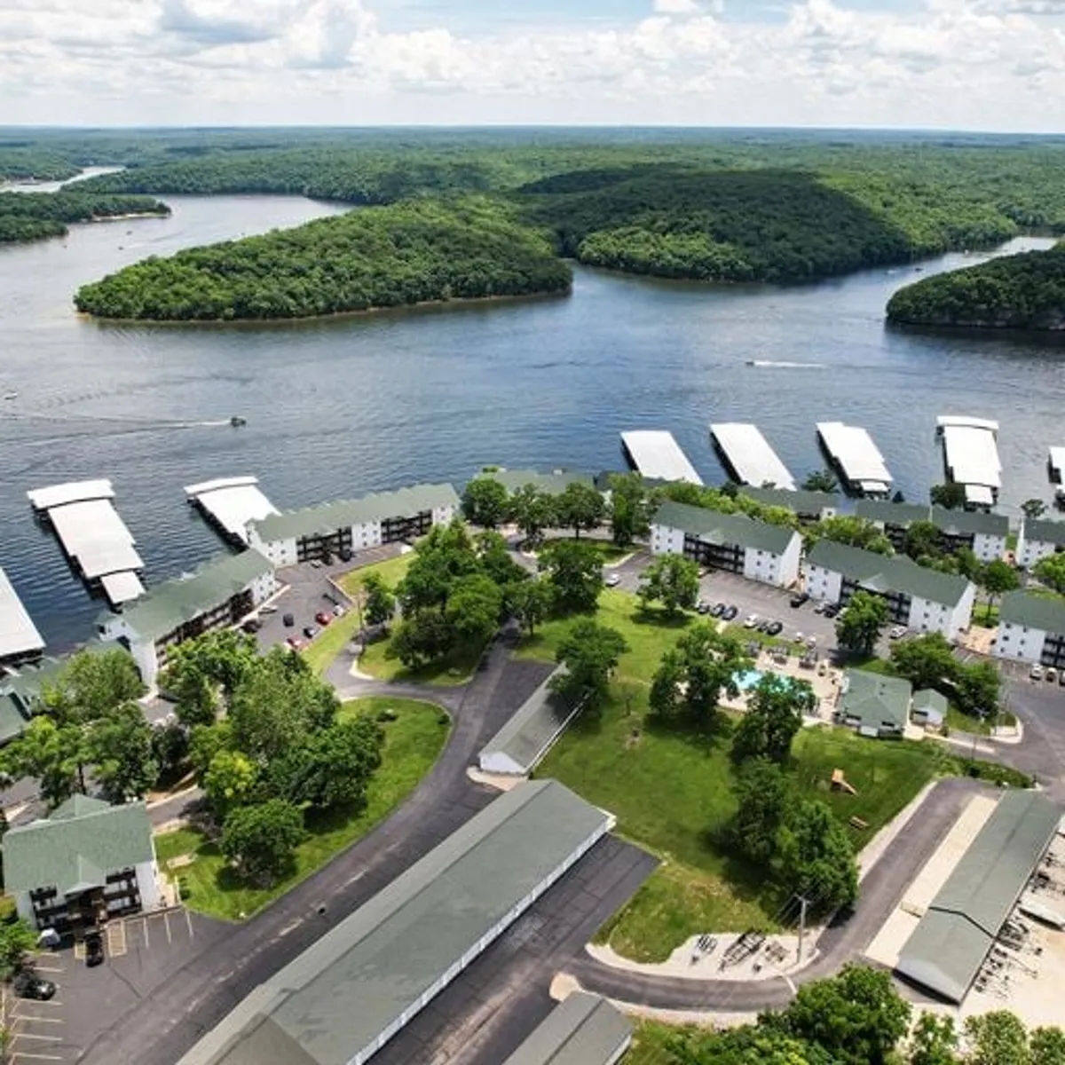 A sweeping exterior shot capturing the lake, green hills, boathouses, residential buildings, and the sparkling swimming pool. The image exudes a sense of relaxation and recreation, with natural beauty and multiple resort-style amenities in one view.