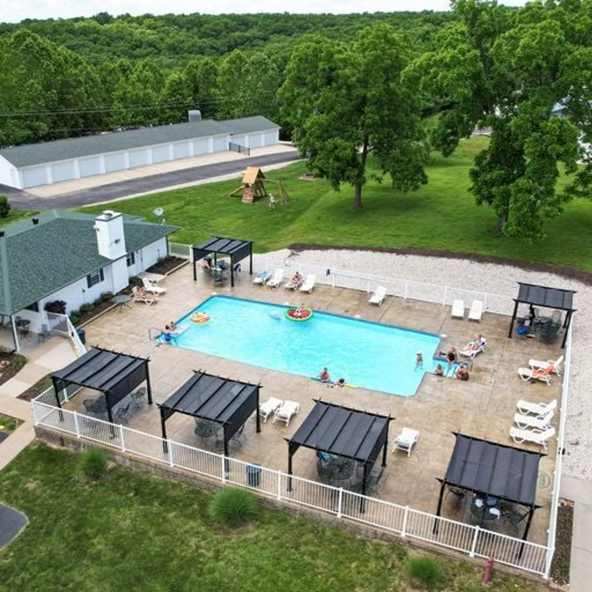 An exterior shot focusing on a playground area with equipment, surrounded by trees, a swimming pool, cabana structures, and lounge chairs. The visible play equipment and cheerful mood reinforce the property’s family-friendly amenities and outdoor spaces for children to enjoy.