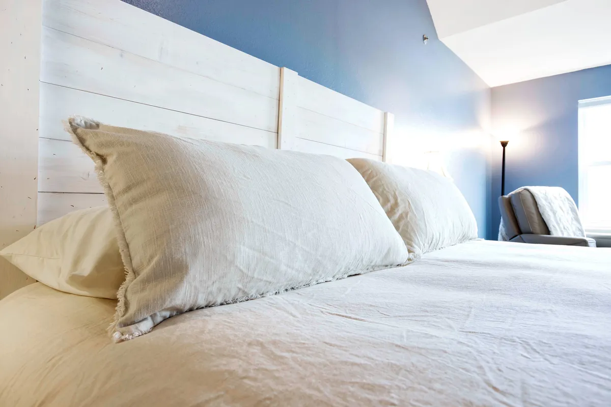 A bedroom scene with large, fluffy pillows on a wooden headboard, soft bedding, a floor lamp, and a window letting in sunlight. The room feels serene and softly illuminated, inviting guests to relax, nap, or wake up gently to natural light.